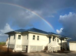 A landscaped, oceanside house with white walls and a blue roof beneath a rainbow